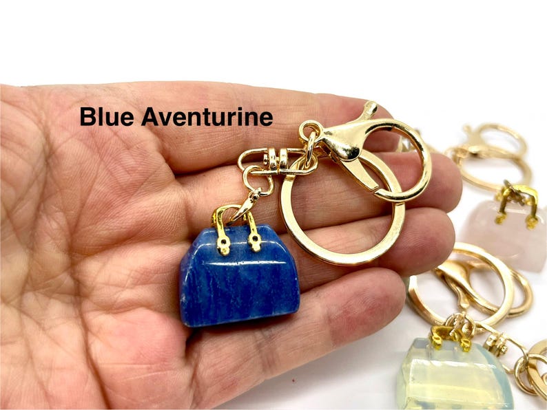 May include: A gold-tone keychain with a blue aventurine handbag charm. The charm is a small, detailed replica of a handbag with gold-tone accents. The keychain is attached to a gold-tone split ring.