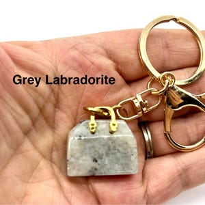 May include: A gold-tone keychain with a grey labradorite handbag charm. The charm is a small, detailed replica of a handbag with a clasp closure. The text "Grey Labradorite" is visible in the image.