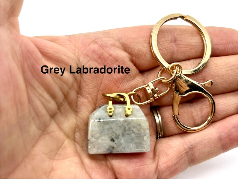 May include: A gold-tone keychain with a grey labradorite handbag charm. The charm is a small, detailed replica of a handbag with a clasp closure. The text "Grey Labradorite" is visible in the image.