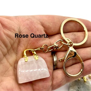 May include: A gold-tone handbag keychain with a rose quartz charm. The keychain has a lobster clasp and a large ring. The text "Rose Quartz" is visible in the image.