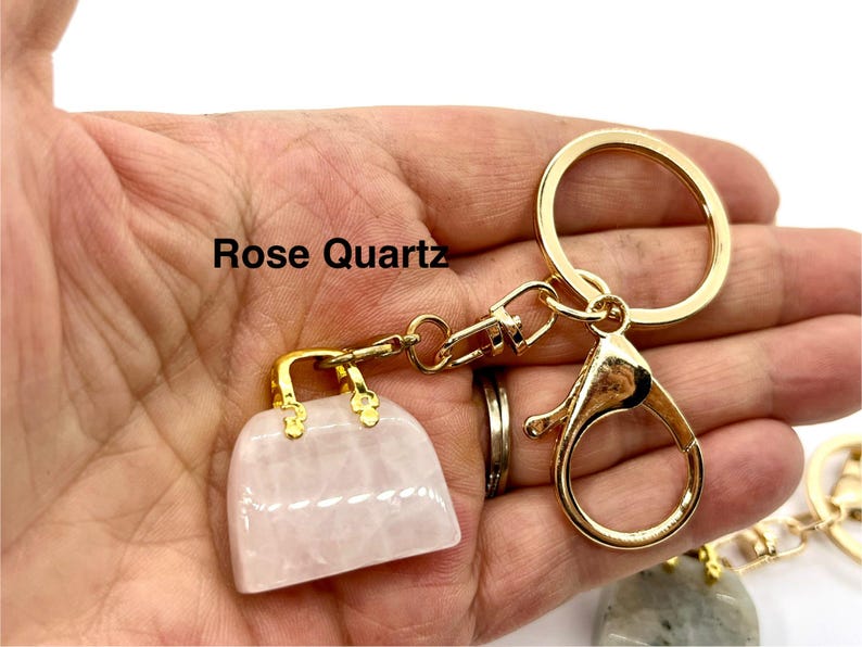 May include: A gold-tone handbag keychain with a rose quartz charm. The keychain has a lobster clasp and a large ring. The text "Rose Quartz" is visible in the image.