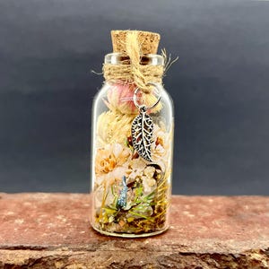 May include: A small glass bottle with a cork stopper filled with dried flowers and greenery. A silver charm in the shape of a leaf hangs from the bottle's neck.