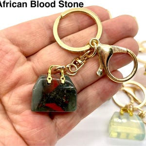 May include: Gold keychain with a small handbag charm made of African Bloodstone. The stone is a dark green with red and black inclusions.