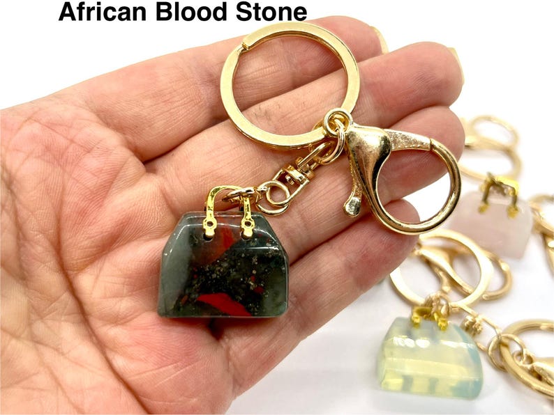 May include: Gold keychain with a small handbag charm made of African Bloodstone. The stone is a dark green with red and black inclusions.