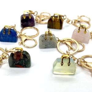 May include: A set of eight gemstone handbag keychains. Each keychain features a different gemstone, including amethyst, tiger's eye, rose quartz, labradorite, jasper, and clear quartz. The keychains are attached to gold-colored key rings.