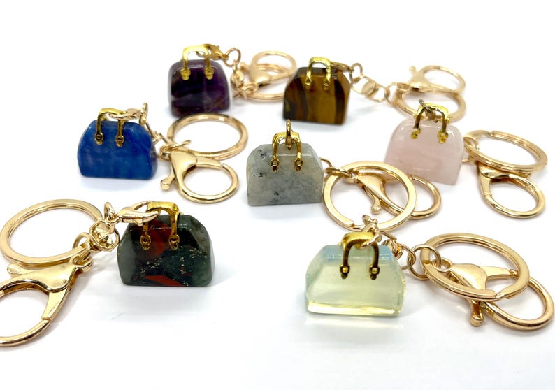 May include: A set of eight gemstone handbag keychains. Each keychain features a different gemstone, including amethyst, tiger's eye, rose quartz, labradorite, jasper, and clear quartz. The keychains are attached to gold-colored key rings.