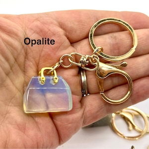 May include: A gold-tone keychain with a clear, iridescent opalite handbag charm. The charm has a gold-tone clasp and handle. The keychain also includes a gold-tone split ring and a lobster clasp.