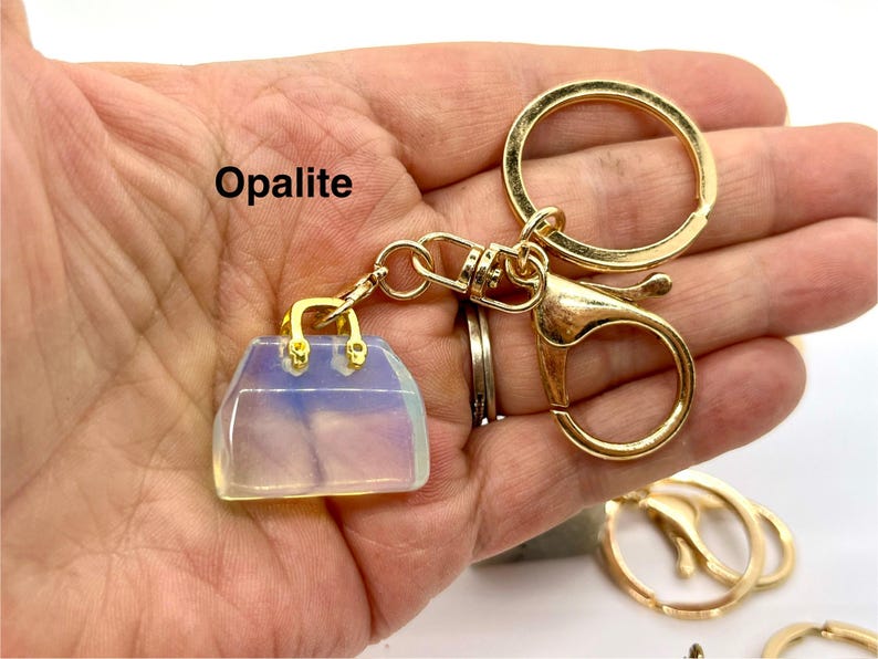 May include: A gold-tone keychain with a clear, iridescent opalite handbag charm. The charm has a gold-tone clasp and handle. The keychain also includes a gold-tone split ring and a lobster clasp.