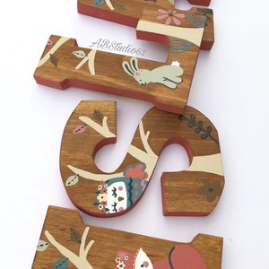 May include: Wooden letters spelling "A-S-I" decorated with a woodland theme. The letters are painted brown with a red border. Each letter features a different woodland animal, including a bear, a rabbit, an owl, and a fox. The animals are painted in a whimsical style with bright colors.