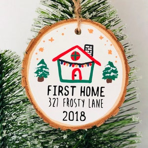 May include: A white wooden ornament with a hand-painted green house with a red roof and a string of lights. The ornament also has two small green trees and the text "FIRST HOME 321 FROSTY LANE 2018".