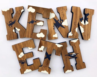 May include: Wooden letters spelling out the name "KENETH" in brown with white clouds and blue airplanes painted on them.