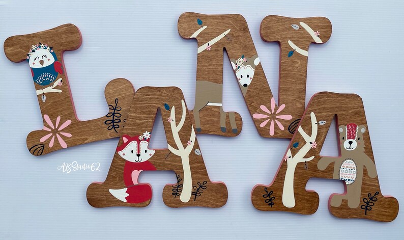 May include: Wooden letters spelling "LANA" with a woodland theme. Each letter is painted brown and features a different animal, including an owl, fox, deer, and bear. The letters are decorated with pink flowers, leaves, and branches.
