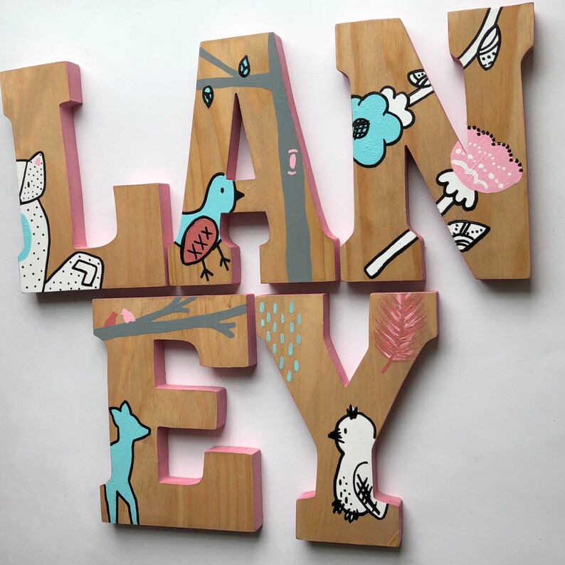 May include: Wooden letters spelling the name "LANEY" painted in pink, blue, and white with nature-themed illustrations. The letters are freestanding and have a rustic, handmade look.