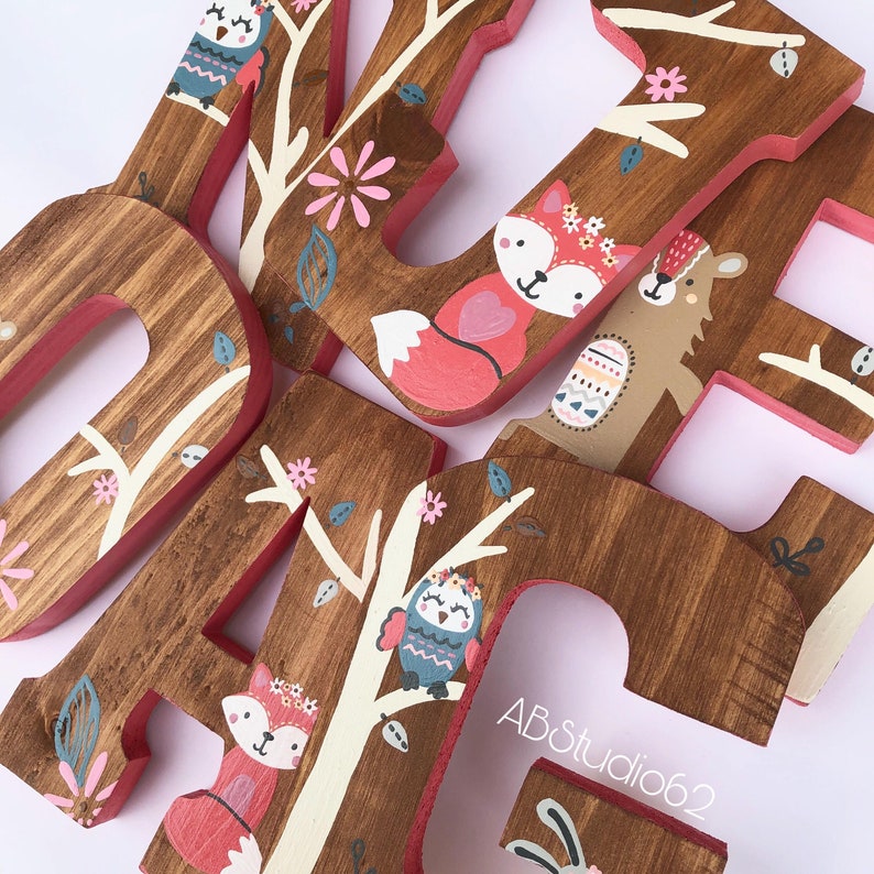 May include: Wooden letters painted with a woodland theme. The letters are painted in shades of brown, pink, and white. The letters are decorated with painted images of owls, foxes, and bears. The letters are arranged in the order "LOVE".