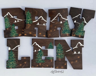 May include: Wooden letters spelling out the name "Braylen" with a woodland theme. Each letter is painted brown and features white mountains, green pine trees, and brown paw prints.