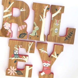 May include: Wooden letters spelling "RILEY" painted in brown with a woodland theme. Each letter features a different animal, including a fox, a bear, an owl, and a rabbit. The letters are decorated with white branches, pink flowers, and green leaves.