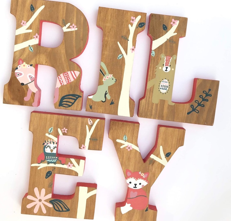 May include: Wooden letters spelling "RILEY" painted in brown with a woodland theme. Each letter features a different animal, including a fox, a bear, an owl, and a rabbit. The letters are decorated with white branches, pink flowers, and green leaves.