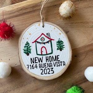 May include: A white wooden ornament with a hand-painted house, trees, and stars. The ornament says "NEW HOME 7164 BUENA VISTA 2023".