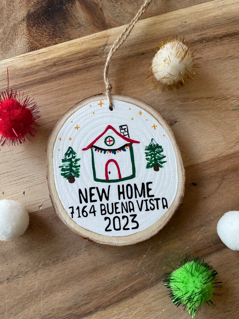 May include: A white wooden ornament with a hand-painted house, trees, and stars. The ornament says "NEW HOME 7164 BUENA VISTA 2023".