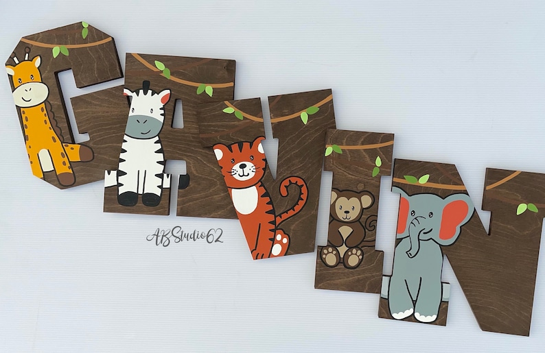 May include: Wooden letters spelling the name "Gavin" with a jungle theme. Each letter is painted brown and features a different animal: a giraffe, zebra, tiger, monkey, and elephant. The letters are decorated with green leaves and vines.