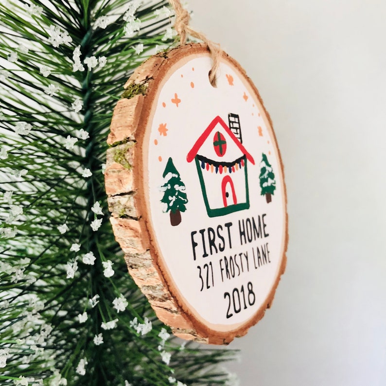 May include: A round wooden ornament with a white background and a hand-painted illustration of a house with a red roof, green trim, and a string of lights. The text "FIRST HOME 321 FROSTY LANE 2018" is written below the house.