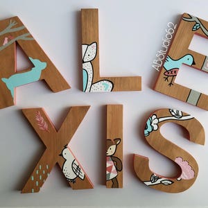 May include: Wooden letters spelling out the name "ALEXIS" painted in various colors with different animal and nature-themed designs. The letters are painted in shades of brown, pink, blue, and white.