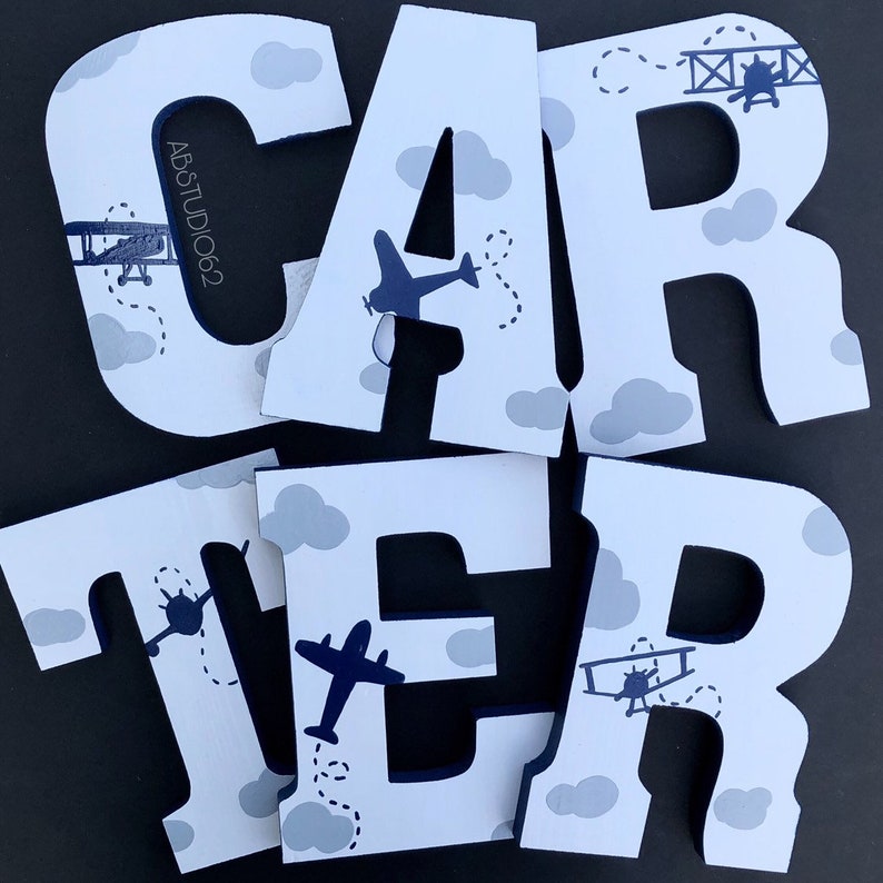 May include: White wooden letters spelling out the name "CARTER" with a blue airplane and cloud design. The letters are painted white with blue accents.
