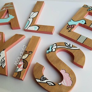 May include: Wooden letters painted with a whimsical woodland theme. The letters are painted in shades of brown, pink, blue, and white. The letters are decorated with images of birds, deer, and flowers. The letters spell out the name "ALISE".