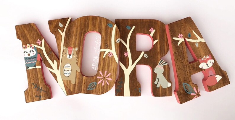 May include: Wooden letters spelling the name "NORA" painted in brown with pink accents. Each letter features a different woodland animal, including an owl, bear, bunny, and fox.
