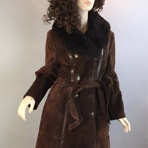 May include: A long, brown suede coat with a black fur collar and a matching belt. The coat has a double-breasted design with gold buttons and leather trim. The mannequin is wearing blue jeans.