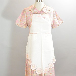 May include: A white apron with pink floral embroidery and the word "MEXICO" stitched on the front. The apron is worn over a pink and white plaid shirt.