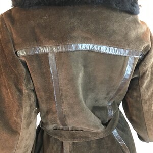 May include: A brown suede coat with a black faux fur collar. The coat has a belt and leather trim detailing. The back of the coat is visible, showcasing the tailored design and texture of the suede material.