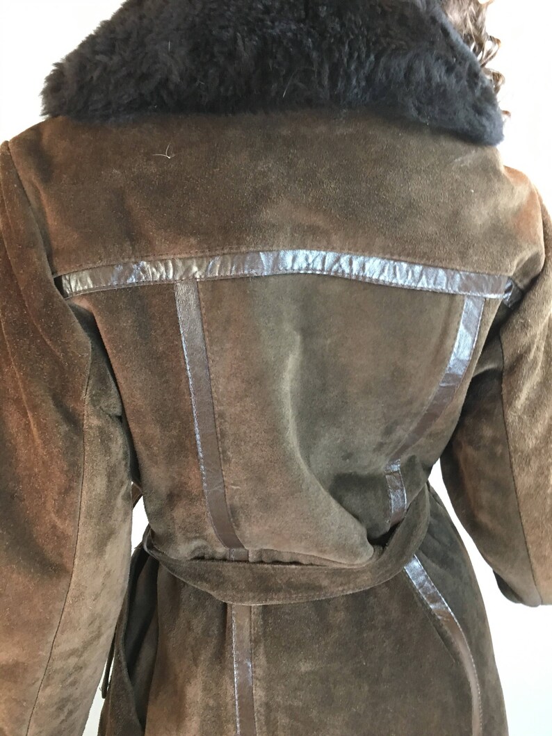 May include: A brown suede coat with a black faux fur collar. The coat has a belt and leather trim detailing. The back of the coat is visible, showcasing the tailored design and texture of the suede material.