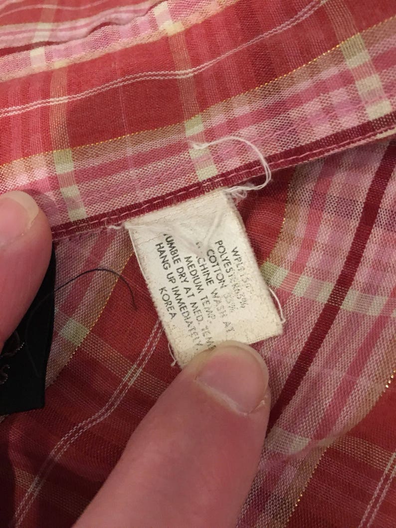 May include: A white care label with black text on a red and white plaid fabric. The text reads: "WPL150 POLYESTER 63% COTTON 33% MACHINE WASH AT MEDIUM TEMP. TUMBLE DRY AT MED. TEMP. HANG UP IMMEDIATELY. KOREA"