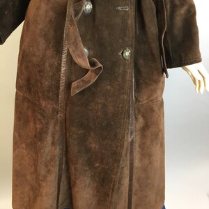 May include: A brown suede coat with a double-breasted design and a matching belt. The coat features large, round buttons and a classic trench coat style. The suede has a textured appearance, and the coat is knee-length.