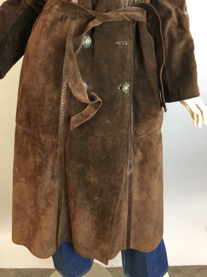 May include: A brown suede coat with a double-breasted design and a matching belt. The coat features large, round buttons and a classic trench coat style. The suede has a textured appearance, and the coat is knee-length.