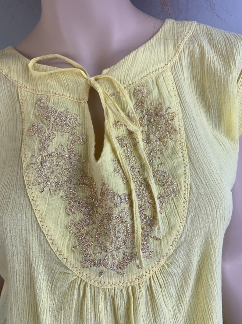 May include: A yellow cotton blouse with a keyhole neckline and gold embroidery. The blouse has a tie at the neckline.
