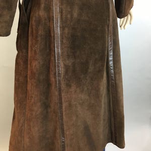 May include: A long, brown suede coat with leather trim. The coat has a wrap-around design and long sleeves. The suede has a soft, textured appearance. The coat is shown on a mannequin, with blue jeans visible at the bottom.