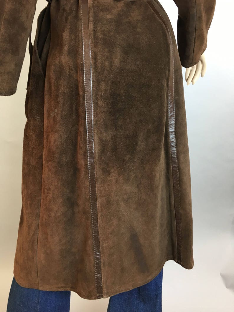 May include: A long, brown suede coat with leather trim. The coat has a wrap-around design and long sleeves. The suede has a soft, textured appearance. The coat is shown on a mannequin, with blue jeans visible at the bottom.