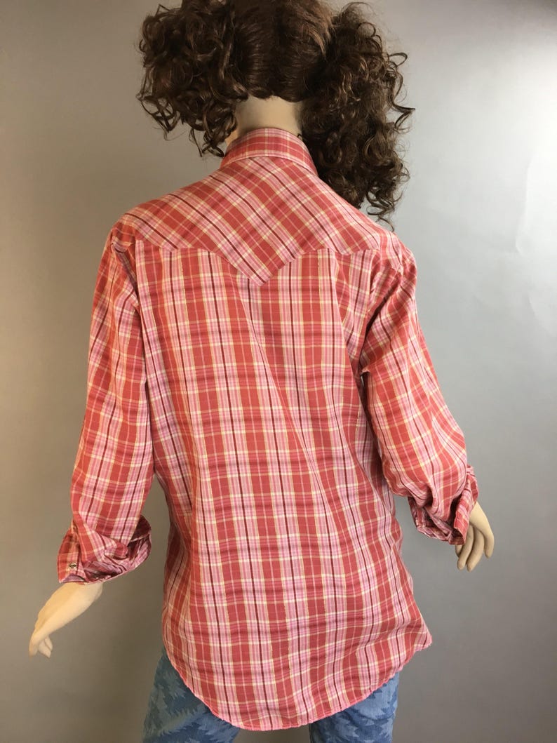 May include: A pink and white plaid long-sleeve shirt with a pointed collar and button closure. The shirt has a yoke in the back and a single button on each cuff.