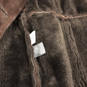 May include: Close-up of a brown shearling coat interior. The coat's lining is a dark brown, plush material. A tag is visible, with text indicating the materials used in the coat's construction. The coat's exterior is a darker shade of brown.