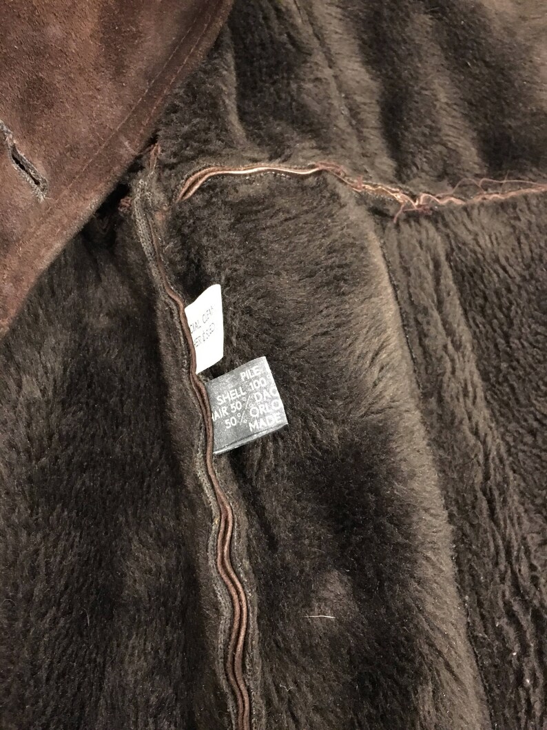 May include: Close-up of a brown shearling coat interior. The coat's lining is a dark brown, plush material. A tag is visible, with text indicating the materials used in the coat's construction. The coat's exterior is a darker shade of brown.