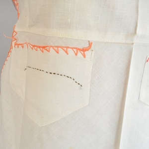May include: White linen dress with two pockets on the back. The pockets are outlined with pink stitching and have a black dashed line stitched across the pocket.