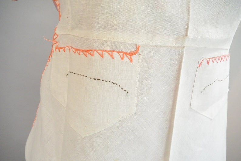 May include: White linen dress with two pockets on the back. The pockets are outlined with pink stitching and have a black dashed line stitched across the pocket.