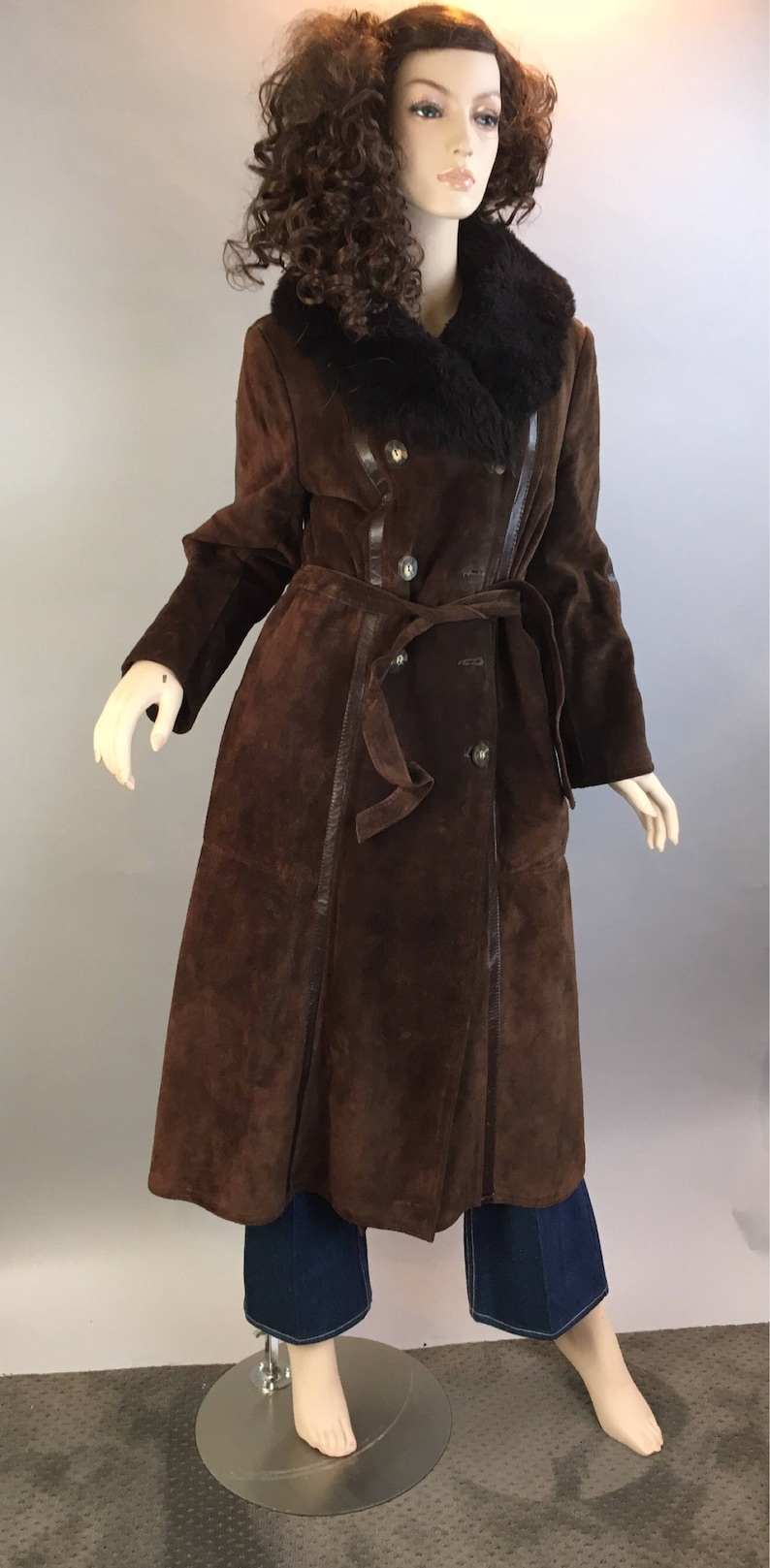May include: A long, brown suede coat with a black fur collar and a matching belt. The coat has a double-breasted design with gold buttons and leather trim. The mannequin is wearing blue jeans.