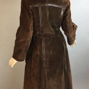 May include: A long, brown suede coat with a dark fur collar and a matching belt. The coat features leather trim and a classic design. The coat is displayed on a mannequin.