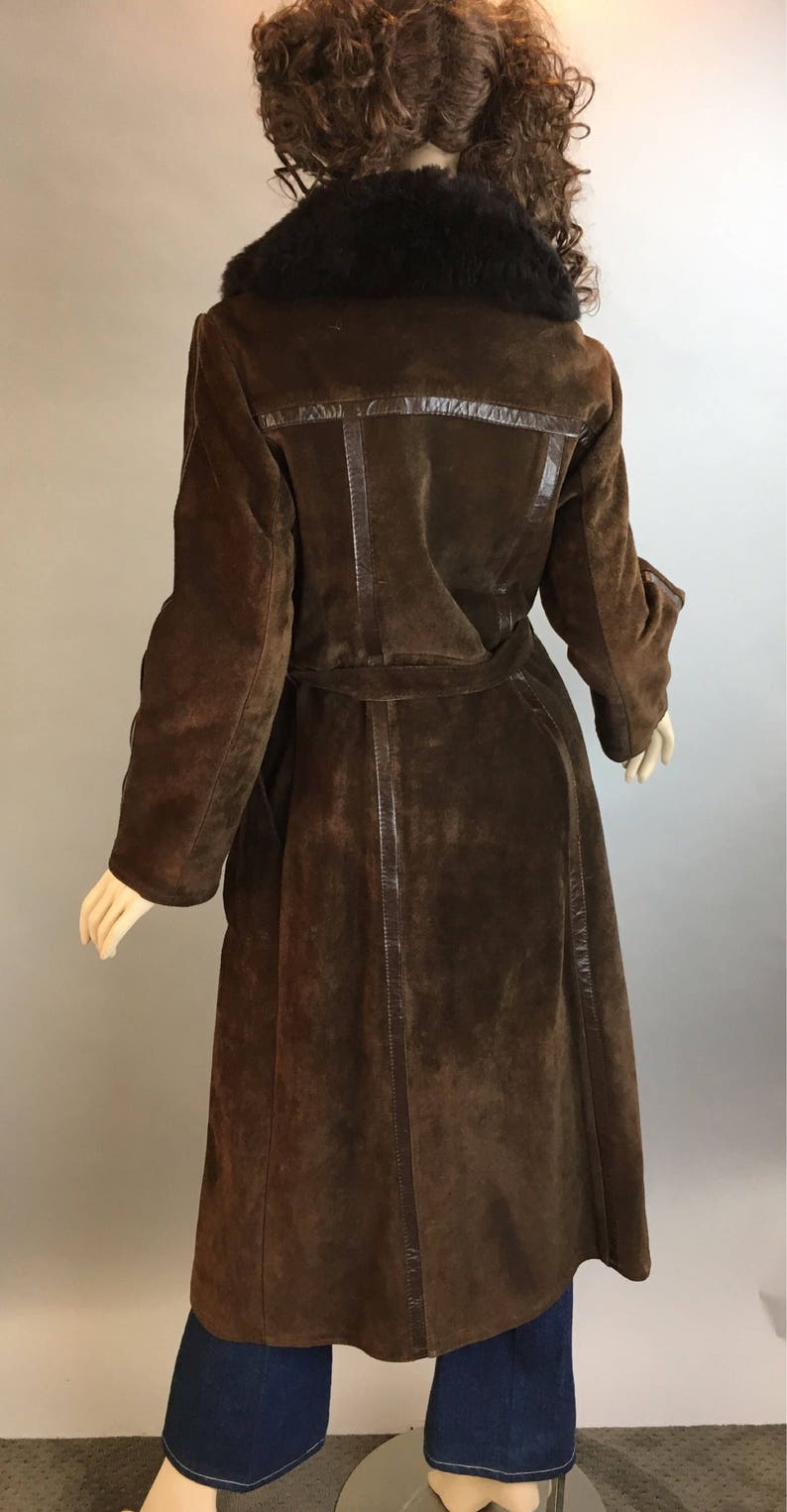 May include: A long, brown suede coat with a dark fur collar and a matching belt. The coat features leather trim and a classic design. The coat is displayed on a mannequin.