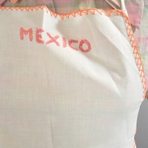 May include: White linen apron with a pink embroidered border and the word "MEXICO" stitched in pink on the front.