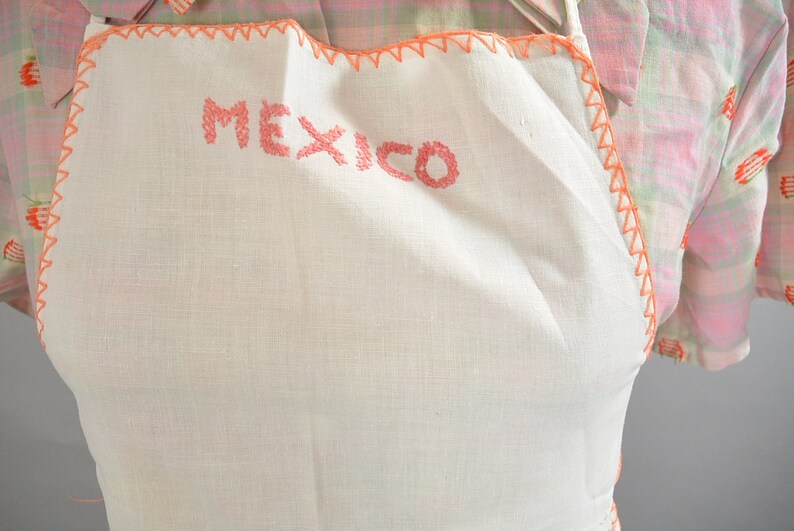May include: White linen apron with a pink embroidered border and the word "MEXICO" stitched in pink on the front.
