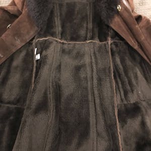 May include: A brown suede coat with a dark brown fur collar and lining. The coat features a button closure and a classic design, ideal for cold weather. The interior is a plush, dark brown material.